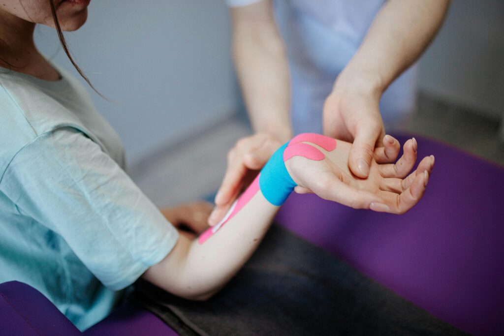 Physiotherapie‑Behandlung am Arm mit farbigem Kinesio‑Tape, Therapeut unterstützt Patientin bei Beschwerden nach Verletzung