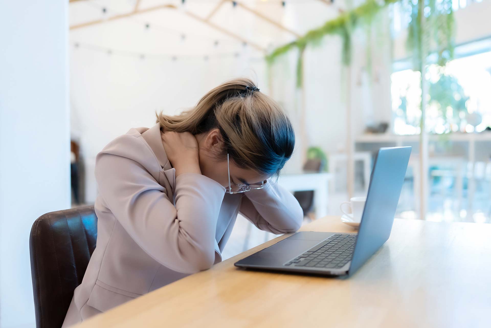 Junge Frau sitzt gebäugt im Büro und hält sich ihren Nacken, zeichen für Nackenverspannung und Schmerzen